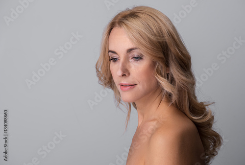 Enigmatic sensual naked middle-aged woman is standing and looking down distantly. Isolated background with copy space in the left side
