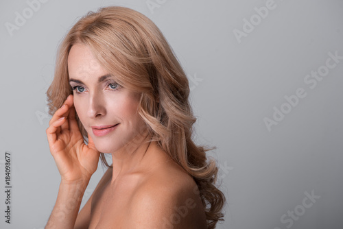 Mysterious beauty. Sensual elegant middle-aged naked woman is standing and touching her face while looking aside thoughtfully. Isolated background with copy space in the right side