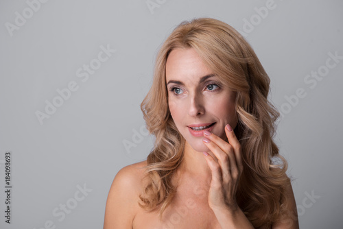 Portrait of delighted charming middle-aged naked woman is touching her lips and opening mouth while looking aside wistfully. Isolated background with copy space in the left side