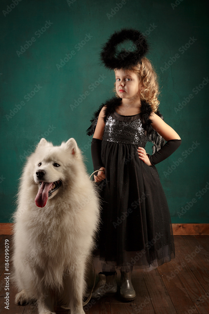 New Year's composition in the studio. Little cute blonde in a black angel costume with a big white dog. Year of the dog, Mood New Year's composition in the studio. Little cute blonde in a black angel costume with a big white dog. Year of the dog, Mood