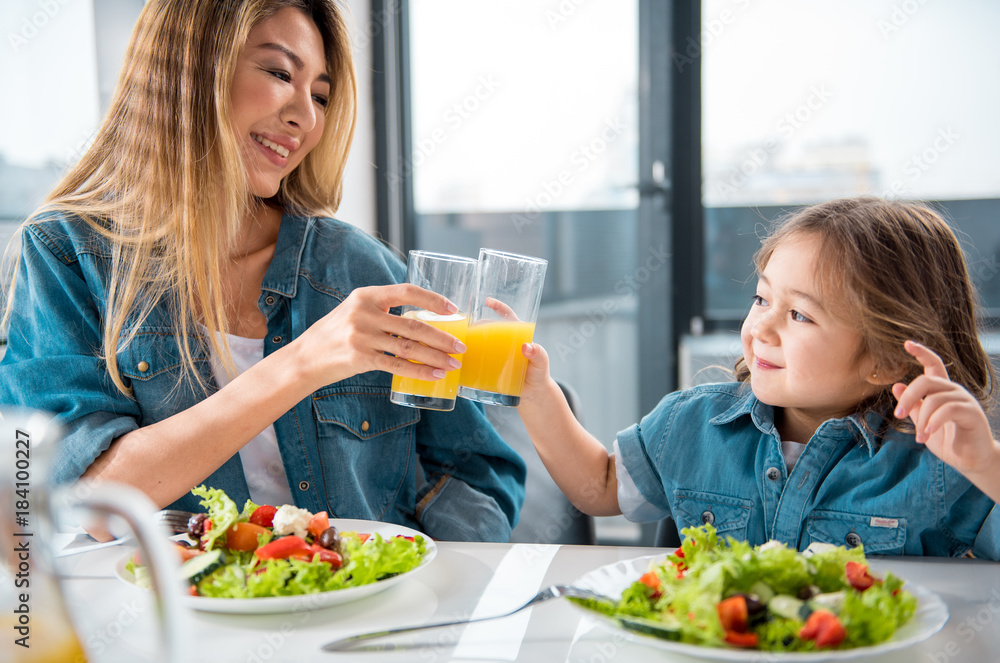 Portrait of happy asian woman and girl clinking glasses of juice and laughing. They are sitting at table and eating salad in kitchen Portrait of happy asian woman and girl clinking glasses of juice and laughing. They are sitting at table and eating salad in kitchen