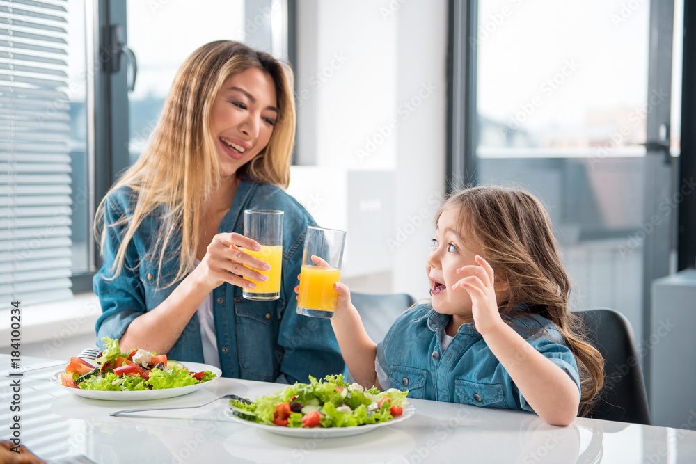 Joyful mother and daughter are enjoying healthy fruit drink. They are looking at each other and smiling while sitting at table