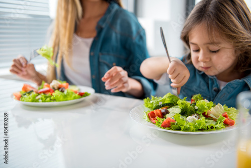 Cheerful mother and daughter are eating healthy salad together at home. Focus on plate with chopped vegetables