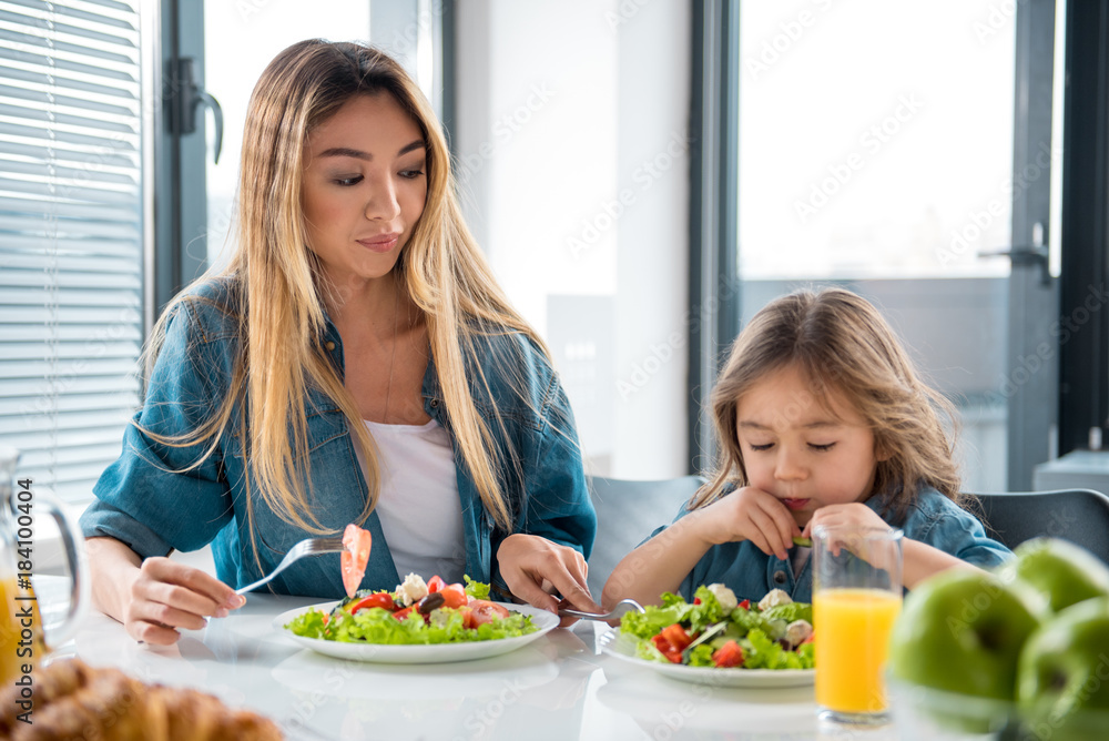Pretty girl is looking at salad in plate with unwillingness. Her mother is giving fork to her while sitting at table in kitchen