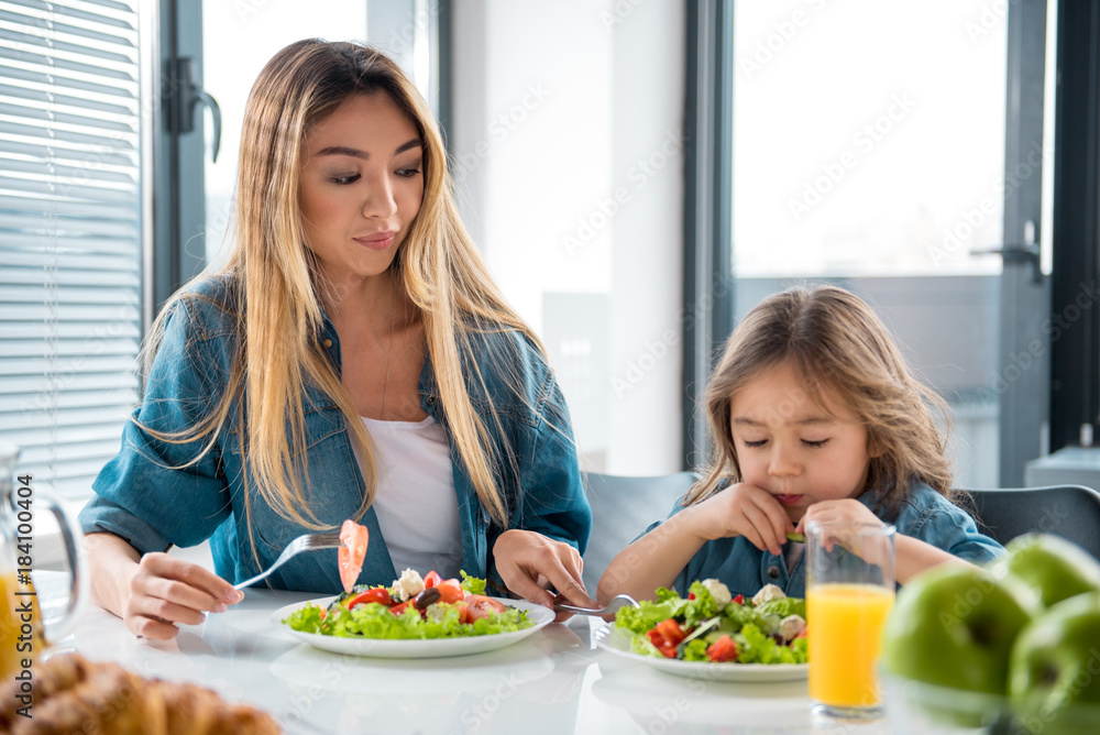Pretty girl is looking at salad in plate with unwillingness. Her mother is giving fork to her while sitting at table in kitchen Pretty girl is looking at salad in plate with unwillingness. Her mother is giving fork to her while sitting at table in kitchen