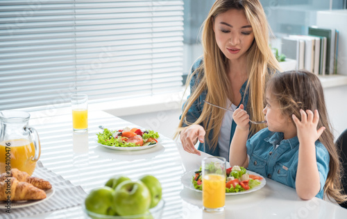 Careful mother is having healthy breakfast with her little daughter. Kid is sitting at table and tasting chopped vegetables with enjoyment. Copy space