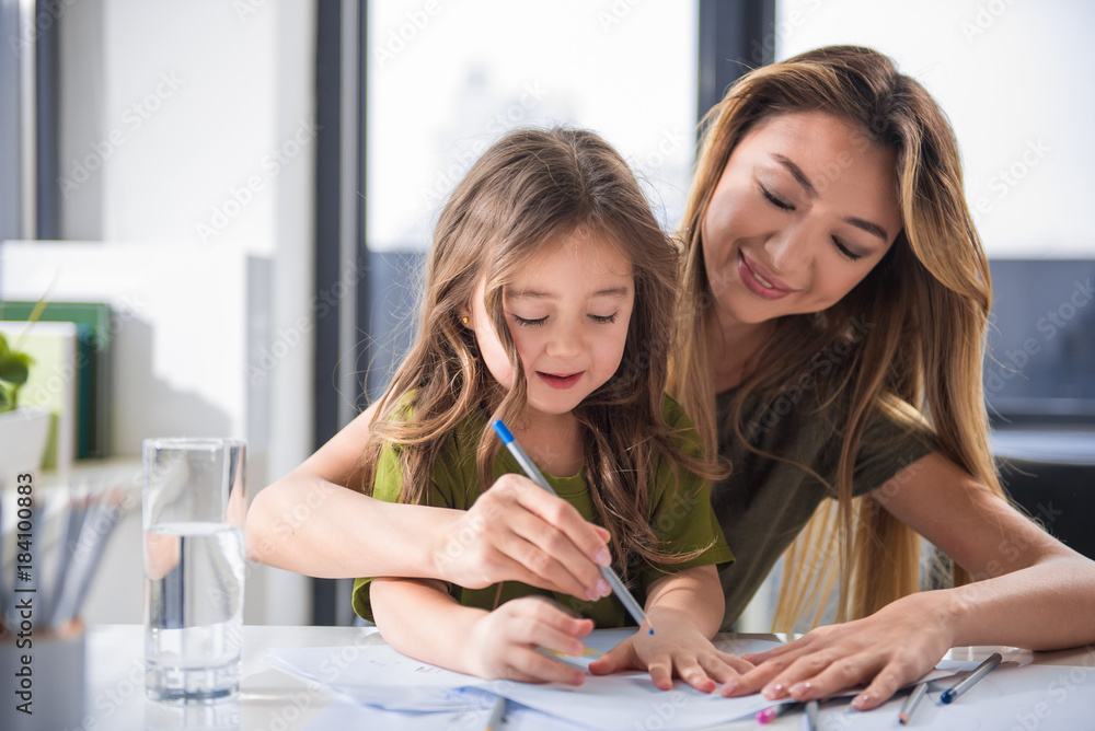 Wait up portrait of joyful mother and daughter drawing image together. They are sitting at home and laughing Wait up portrait of joyful mother and daughter drawing image together. They are sitting at home and laughing
