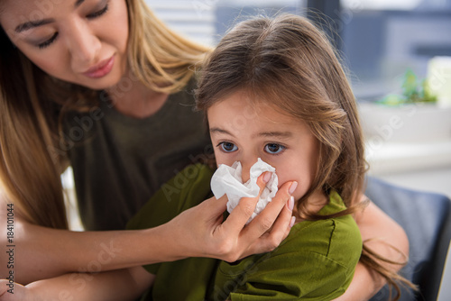 Portrait of cut asian girl blowing snot into the napkin. Her mother is keeping it near her nose with care