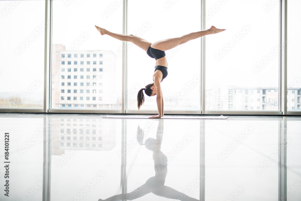 Young attractive yogi woman practicing yoga concept, standing in variation of Pincha Mayurasana exercise, handstand pose, working out, wearing sportswear, full length against windows