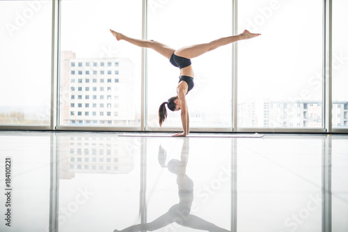 Young attractive yogi woman practicing yoga concept, standing in variation of Pincha Mayurasana exercise, handstand pose, working out, wearing sportswear, full length against windows