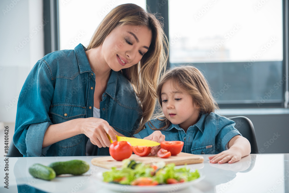 Waist up portrait of joyful asian mother is teaching her child how to make a salad. Girl is looking at her hands with interest Waist up portrait of joyful asian mother is teaching her child how to make a salad. Girl is looking at her hands with interest