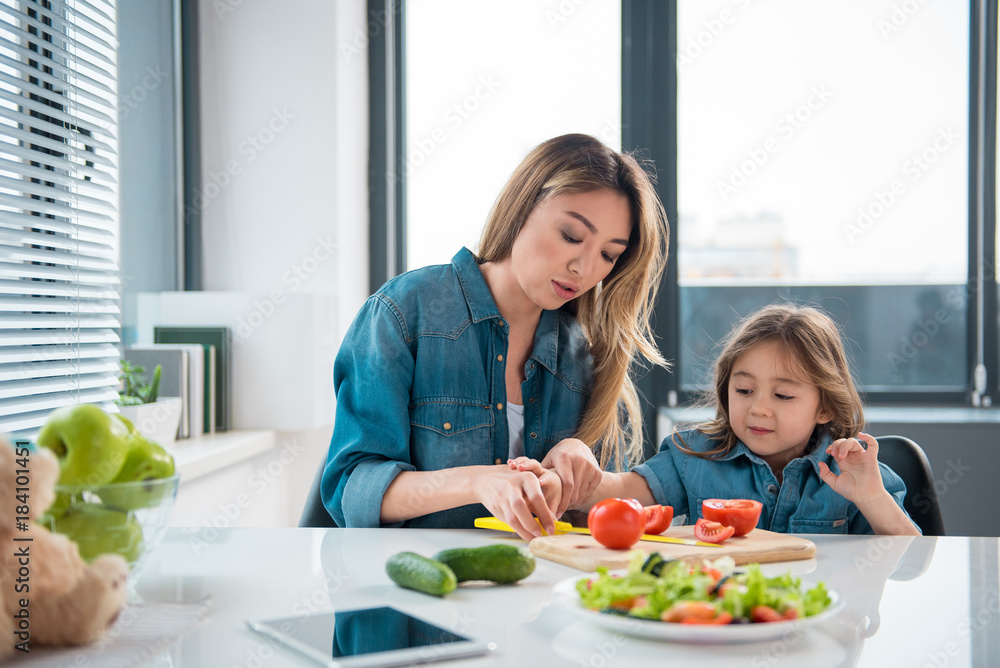 Let me try. Pretty girl is stretching hand to knife with interest while her mother is preparing salad. They are sitting at table in kitchen