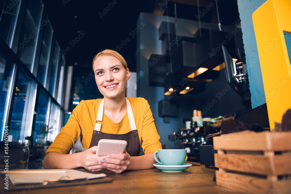 Fototapeta premium Low angle portrait of glad girl barista typing in phone while tasting cup of coffee at table in confectionary shop. Rest concept