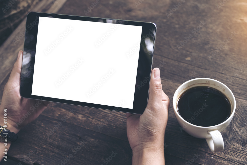 Mockup image of business woman's hands holding black tablet pc with blank white screen and coffee cup on vintage wooden table in cafe background Mockup image of business woman's hands holding black tablet pc with blank white screen and coffee cup on vintage wooden table in cafe background