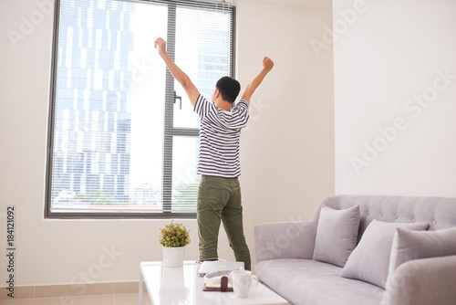 Rear view of a young Asia man stretching his arms near window at home