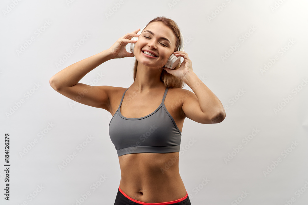 joyful smiling girl in grey sport top with headphones on her head poses for a Studio portrait on a white background with a wide smile and holding hands headphones joyful smiling girl in grey sport top with headphones on her head poses for a Studio portrait on a white background with a wide smile and holding hands headphones