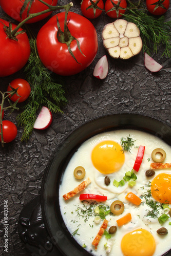 Fried eggs in a cast iron pan with peppers, tomatoes, capers, herbs and bread on a black background