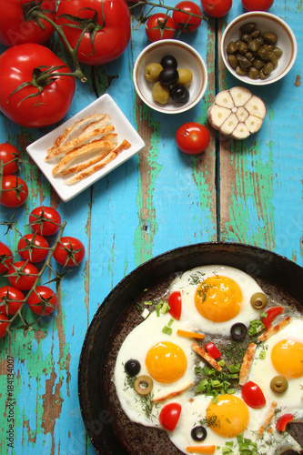 Fried eggs in a cast iron pan with peppers, tomatoes, capers, herbs and bread on the blue boards