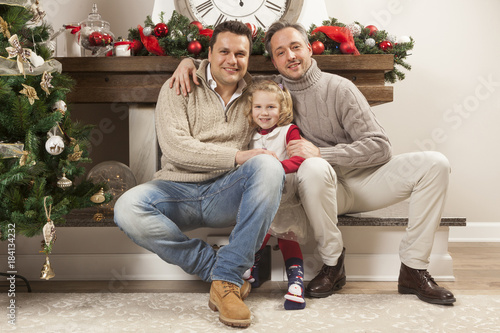 Happy family portrait on Christmas, gay male couple and one child sitting near fireplace at home, chritmas decoration around them © Tommaso Lizzul