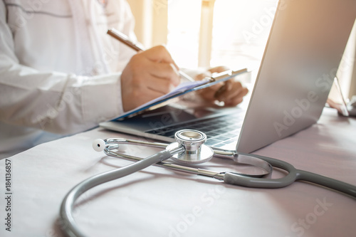 Medicine doctor's writing on laptop in medical office.Focus stethoscope on foreground table in hostpital.Stethoscope is acoustic medical device for auscultation,listening internal sounds of human body photo