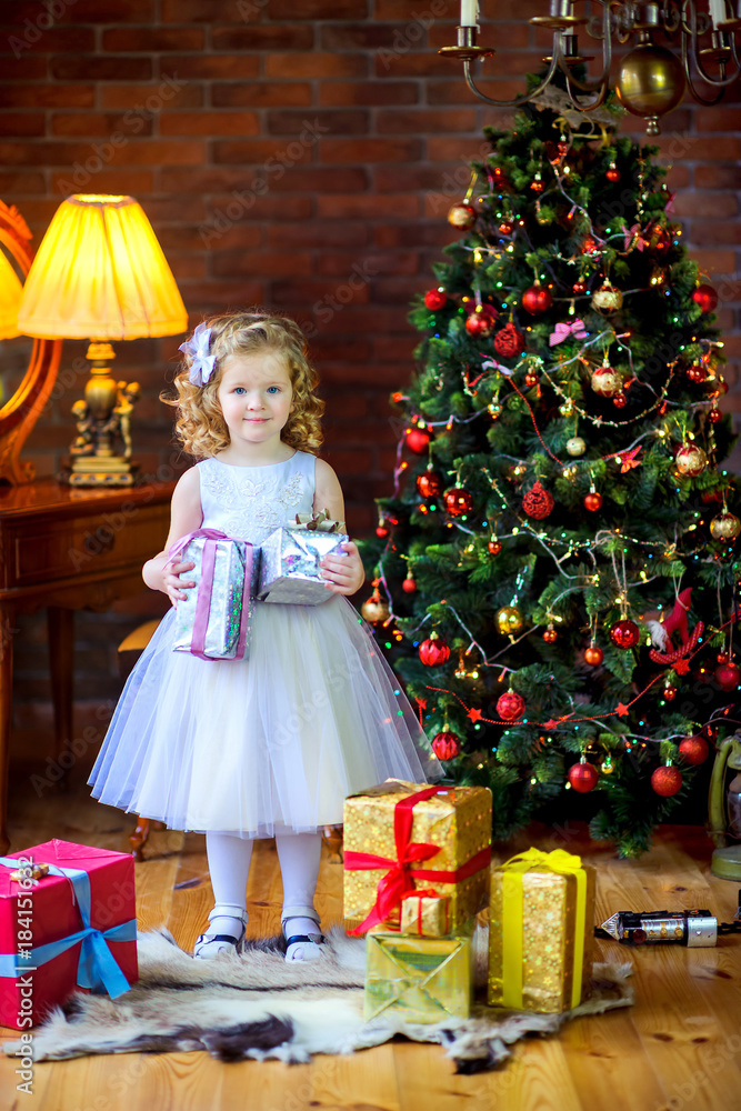 beautiful little i girl in festive dress holding gifts in hands, in the background Christmas tree.