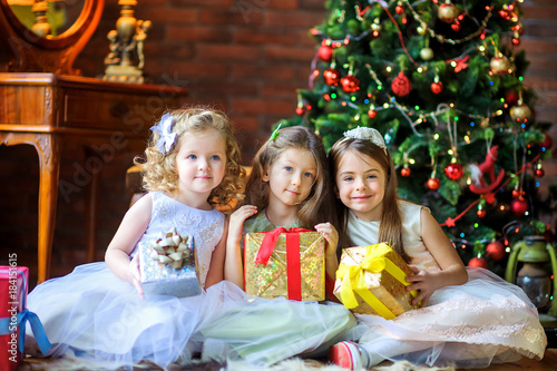 three beautiful girls are sitting on the floor with gifts in hands, in the background a festive Christmas tree.
