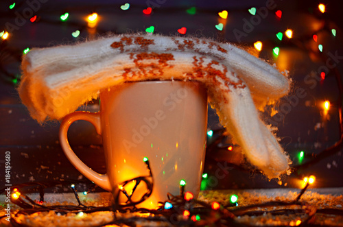 Cup of hot tea or coffee with warm knitted gloves  with garland lights on gray wooden background. Merry Christmas, heart bokeh, snow