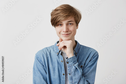 Happiness, positiveness and people concept. Smiling pleased fair-haired young man in denim shirt, keeps hand under chin, rejoices coming weekends, has grandious plans to spend time with friends photo