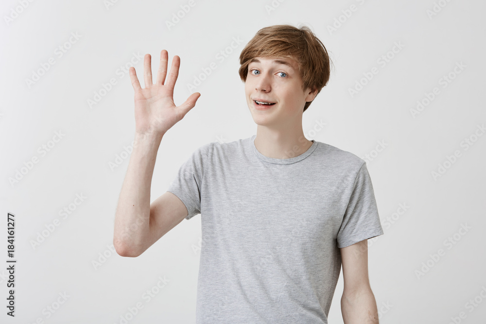 Friendly-looking polite young caucasian man dressed in grey t-shirt smiling, saying hi, waving his hand. Positive human emotions, facial expressions, feelings, attitude and reaction Friendly-looking polite young caucasian man dressed in grey t-shirt smiling, saying hi, waving his hand. Positive human emotions, facial expressions, feelings, attitude and reaction