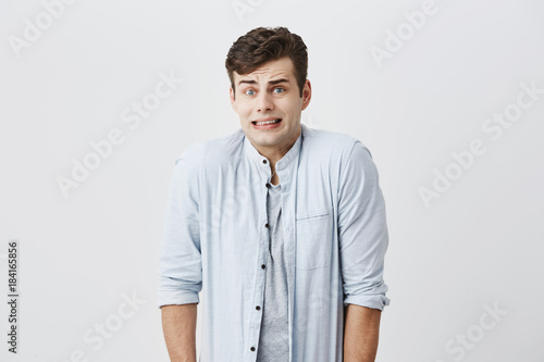 Emotional clueless young male student dressed in blue shirt over gray t-shirt having confused puzzled look, frowning face, showing teeth, shrugging shoulders as he doesn't know reason of accident © Cookie Studio