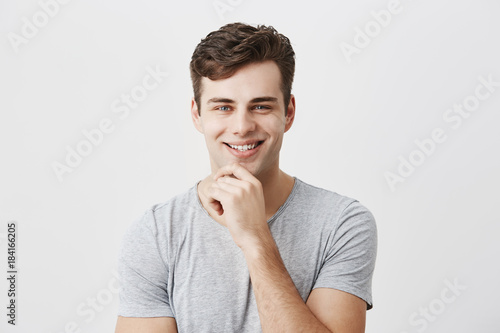 Happy smiling young man demonstrates positive emotions or feelings, has trendy hairdo, dressed casually, keeps hand on chin, stands against gray wall with copy space for text or advertisment.