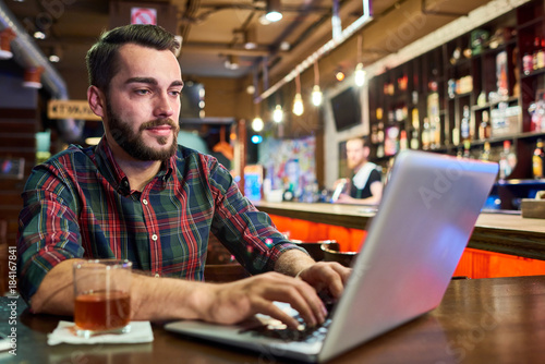 Portrait of modern young man working with laptop sitting at bar counter in beer pub, copy space