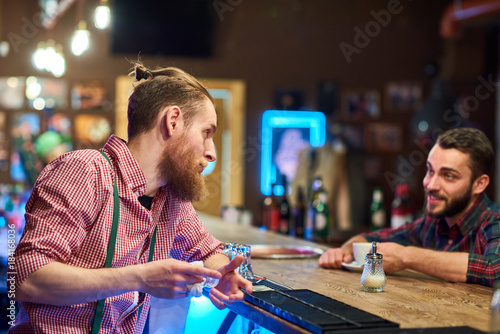 Portrait of modern bearded bartender talking to client at bar counter in pub, copy space