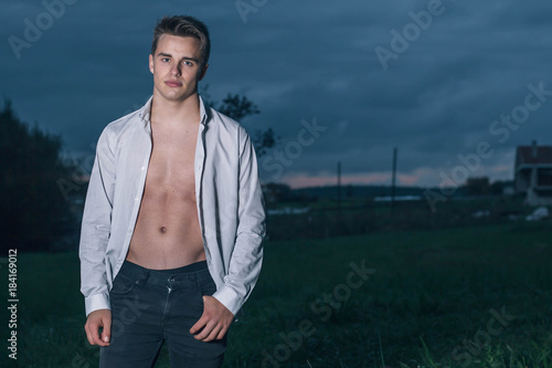 Young model man with open shirt posing standing at dusk in the countryside. © GENETTICA