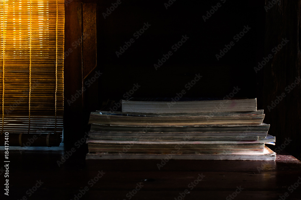 Low key image of stack of books setting on wooden table with sunlight through curtain in the background. (Selective focus)