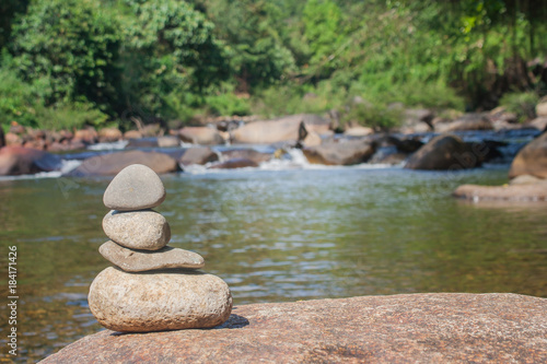 Close up pile of pebbles with beautiful landscape view of small waterfall in the river with water stream flowing through stone and green trees in the background. (Selective focus)