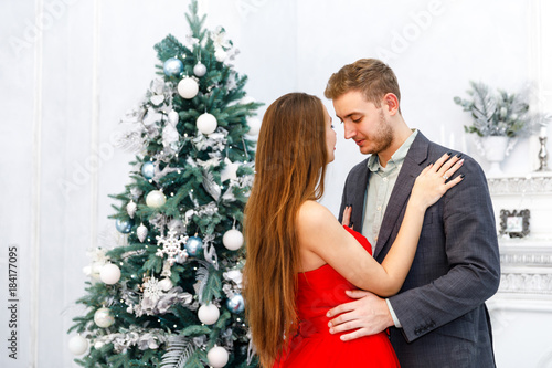 Young smiling couple in evening outfits (jacket and red dress) in white interior with christmas tree © Полина Власова