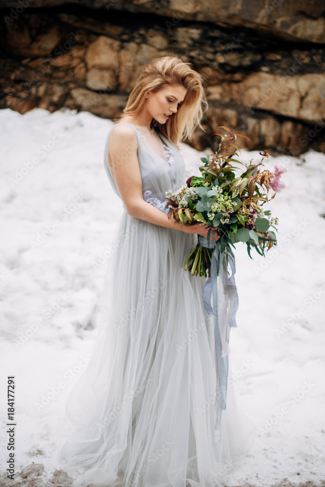 Beautiful woman stands on the road in snowy mountains in grey wedding dress with bouquet in hands Beautiful woman stands on the road in snowy mountains in grey wedding dress with bouquet in hands