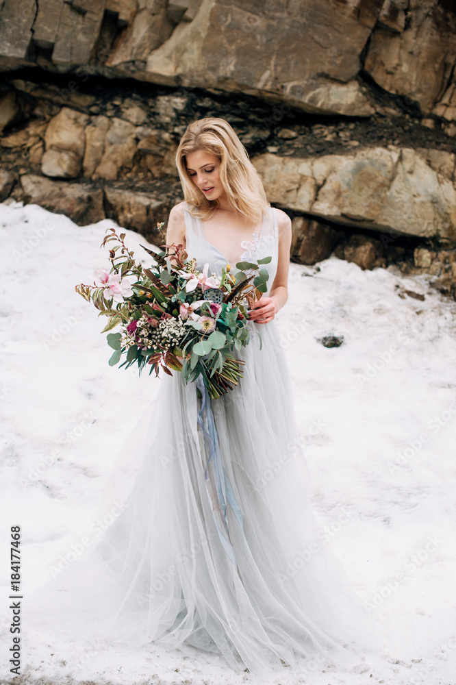Beautiful woman stands on the road in snowy mountains in grey wedding dress with bouquet in hands