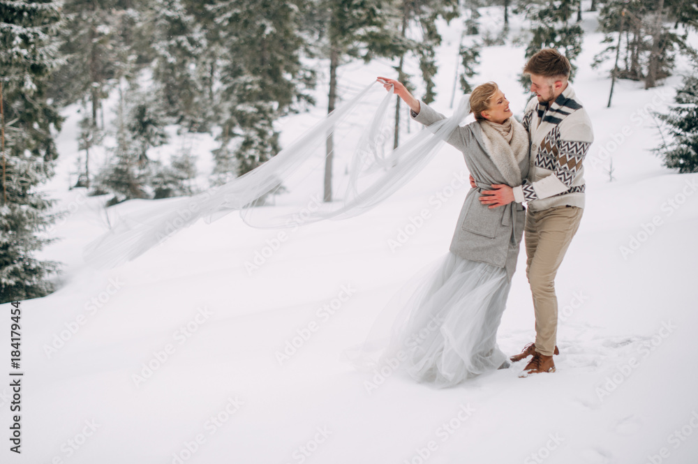 Beautiful couple stands in winter pine forest, woman in grey wedding dress and long veil, bearded man in sweater