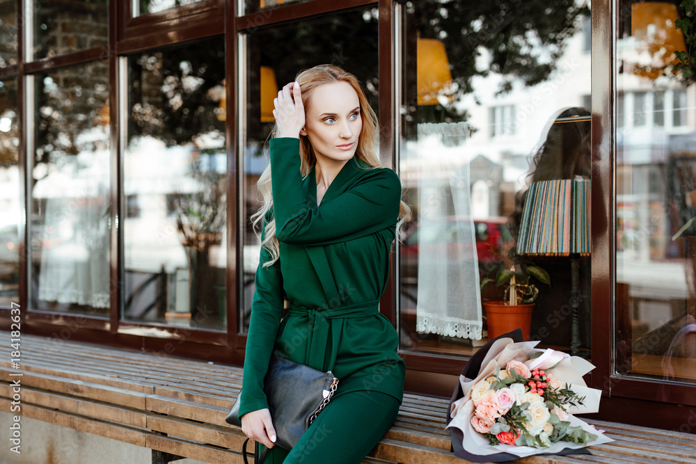 Street style. Portrait of the beautiful young woman of the blonde with a bouquet of flowers in a green suit against the background of city cafe.
