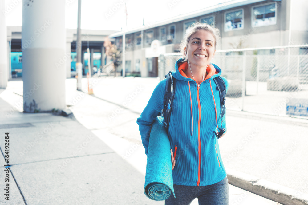 Obraz premium Athlete woman with backpack, headphones, yoga mat going to sport club on the city street. Smiling. Sport tight clothes.