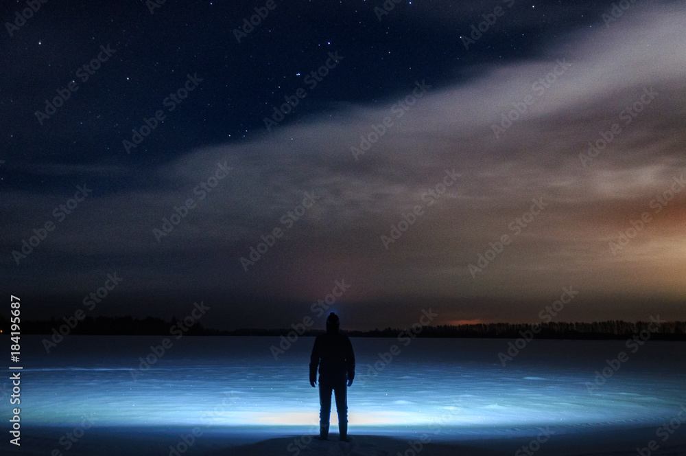 A man with a flashlight on his head is standing and looking at the beautiful starry sky with clouds. In a snowy field. Winter, a frosty night.