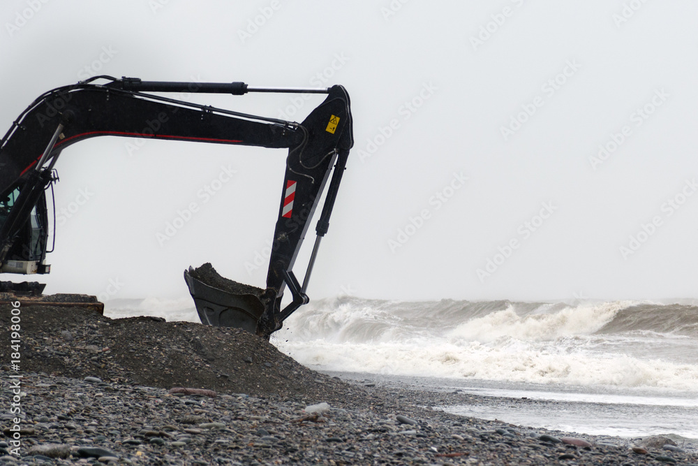 Fototapeta premium The excavator works with a bucket full of stones on the seashore in stormy weather.