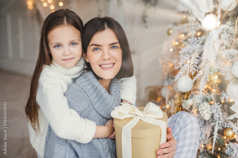 Adorable blue eyed small kid huggs with great love her mother who holds wrapped gift box, stand near decorated New Year tree, happy to celebrate winter holidays. People, celebration, presents concept Adorable blue eyed small kid huggs with great love her mother who holds wrapped gift box, stand near decorated New Year tree, happy to celebrate winter holidays. People, celebration, presents concept