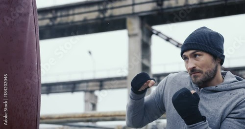 Attractive young sportsman in a hoodie and cap having trainingship of boxing on the cold grey day. Outside the huge old plant ruins. photo