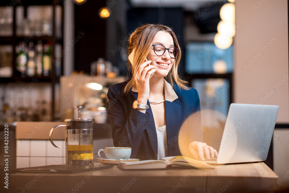 Young businesswoman strictly dressed in the suit talking phone while working with laptop at the modern cafe interior Young businesswoman strictly dressed in the suit talking phone while working with laptop at the modern cafe interior