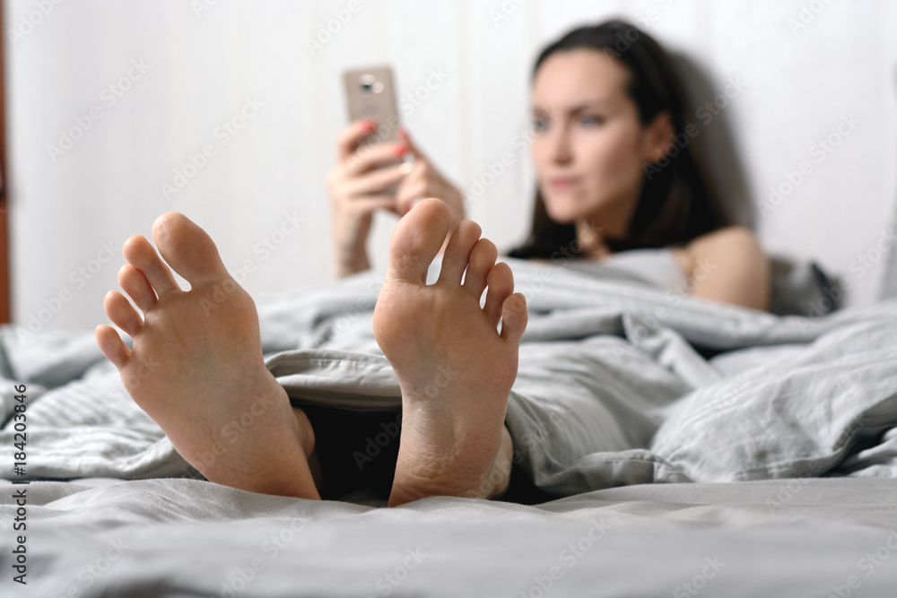 a young woman lies in bed and communicate in social networks via smartphone. focus on the feet. shallow depth of field a young woman lies in bed and communicate in social networks via smartphone. focus on the feet. shallow depth of field
