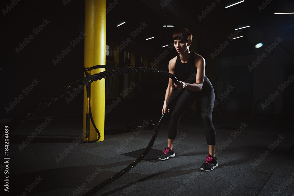 fitness girl doing crossfit exercises in gym with big rope in dark