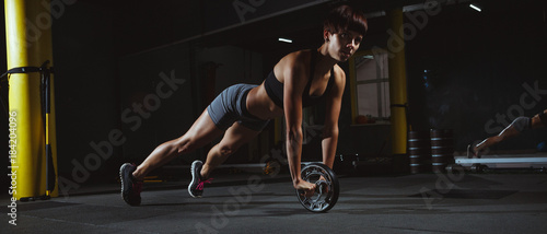fitness girl doing crossfit exercises in gym with wheel in dark hall, sporty woman sexy in pose doing functional training, poster banner healthy lifestyle